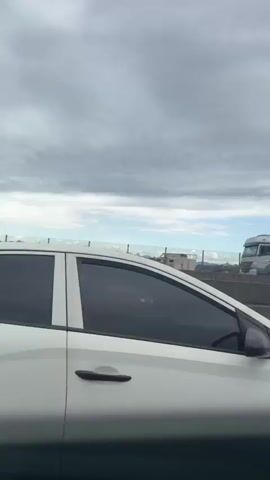 The video shows a view from inside a vehicle driving on a highway. In the foreground, a white car is partially visible. Further ahead, a gray SUV is in the adjacent lane. A large blue shipping container with "COSCO SHIPPING" written on it is being transported by a semi-truck. Beyond the highway, there is a concrete barrier, a chain-link fence, some buildings, and mountains in the distance under a cloudy sky. The camera pans slightly, showing different parts of the highway and the surrounding landscape. At one point, the video briefly focuses on the rear of a red truck with a wooden cargo bed, showing its suspension and tires.