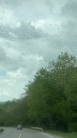 The video shows a view from inside a car driving on a wet road. Raindrops are visible on the windshield, obscuring the view slightly.  Trees line the side of the road, with the leaves appearing bright green. The sky is overcast with grey clouds, with hints of blue visible.  A car is seen driving ahead on the same road.
