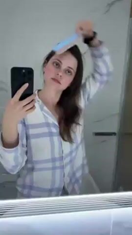 A woman is brushing her hair in front of a bathroom mirror while holding her cell phone and recording herself. She is wearing a blue and white plaid pajama top. The bathroom has a white marble wall. The mirror has a light fixture.