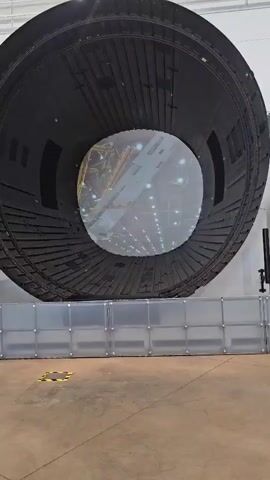A wide-angle shot shows a large, circular, black, concave structure, possibly a piece of an aircraft or a simulator component, tilted at an angle, with its open end facing the camera. Inside this structure, a projection displays an image of what appears to be an aircraft engine and fuselage being worked on by several people in a workshop or factory setting. The projection fills the circular opening, creating a layered visual effect. In the foreground, a low, translucent barrier made of panels and metal frames separates the viewer from the main structure. The ground is a concrete floor with yellow markings. The environment appears to be an indoor industrial space with bright overhead lighting. The video shows a subtle rotation or movement, suggesting the camera is panning or the projected image is changing or moving.