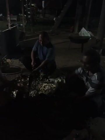 Two people are in a dark, cluttered space. One person, wearing a blue shirt, is squatting and using a tool to chop or break something inside a large container or tub. Another person, wearing a grey shirt and glasses, is also squatting and looking into the same container. There appears to be a collection of what may be chopped materials inside the container. The area is dimly lit, with various objects and structures visible in the background.
