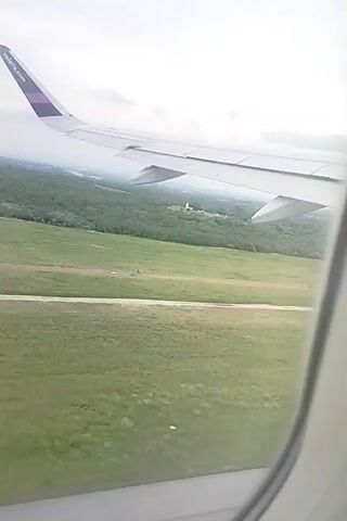The video shows a view from an airplane window during descent or landing. The wing of the plane, marked with "volaris.com," is visible. Below, there is a green landscape consisting of fields, trees, and possibly some water features. A few buildings can be seen among the trees. The sky is overcast, with a gray tone. The overall scene suggests a rural or suburban environment being approached by the aircraft.