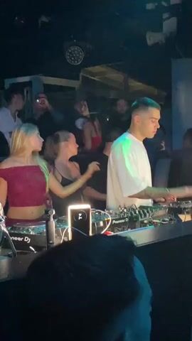 The video shows a DJ in a nightclub playing music. The DJ, a man with short dark hair and tattoos on his arms, is wearing a white t-shirt and standing behind a DJ console with Pioneer DJ equipment. Two women are standing next to him, one with blonde hair wearing a pink top and the other with brown hair in a ponytail wearing a black tank top. The background is dark, with club lighting and other people in the background. The overall atmosphere appears to be a lively party.