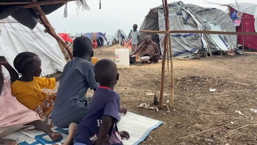 A group of children are seen from behind, sitting on mats on the ground. They appear to be in a makeshift shelter with a thatched roof. In the background, several tents and other shelters are visible, suggesting a camp environment. An adult, possibly a woman, is crouched down near a cooking pot in front of one of the tents. Another adult, a man, stands a short distance away. The ground is dusty and uneven. A large white water jug is placed near the cooking area. The overall scene depicts a humble living situation, likely in a refugee or displacement camp.