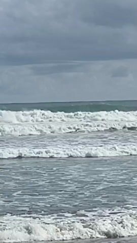 The video shows a choppy ocean with several sets of waves rolling towards the shore. The sky is overcast with dark clouds. The water is a mix of grey and blue. There are no people or objects visible in the video. The waves appear to be of moderate size and are breaking with white foam. The video is shot from a low angle, close to the water's surface. The overall mood of the video is calm and serene, despite the rough sea.