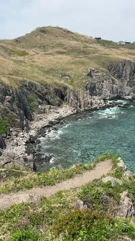 The video shows a coastal landscape with a rocky shoreline meeting a teal-colored sea. The camera pans from a dirt path on a grassy cliff edge to reveal more of the coastline. Further along the cliff, there's a building or structure on a hill. The sea extends to the horizon under a sky that transitions from overcast to brighter blue, while large rock formations stand in the water, battered by the waves.