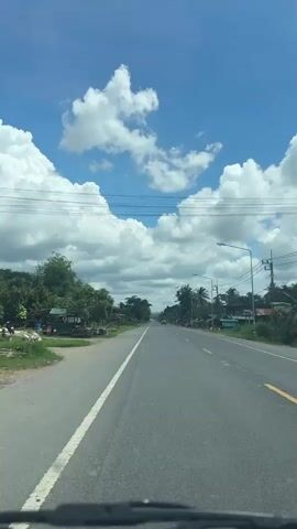 The video shows a view from inside a vehicle driving on a paved road. The sky is blue with large, fluffy white clouds. On either side of the road are trees, vegetation, and small buildings that appear to be houses or shops. There are also utility poles and wires visible along the roadside. A few cars can be seen in the distance on the road. The road has a white line marking the lane and a yellow line on the right side. The perspective suggests the video is captured from the driver's or front passenger's viewpoint.