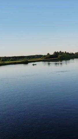 A serene aerial shot captures a vast, calm body of water, likely a river or lake, under a clear, bright blue sky, suggesting a pleasant daytime. The water reflects the sky and the distant treeline, creating a peaceful, almost mirror-like surface. In the middle distance, a small, light-colored boat with at least one person paddling can be seen. To the right, a red and white buoy marks the waterway. The background is a lush green landscape with a dense treeline and scattered vegetation, indicative of a natural, perhaps rural, setting. The overall mood is tranquil and idyllic, evoking a sense of quiet solitude and natural beauty. The timestamp "00:00" appears in the upper left corner, indicating the beginning of the video clip.