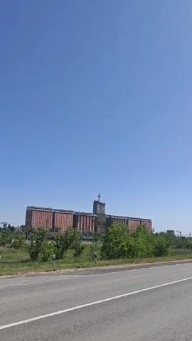 The video shows a grain elevator building viewed from a moving vehicle on a road. The sky is blue. The building is long and appears to be constructed of brick with concrete or similar materials. It features a central tower topped with a communication antenna. The road is asphalt, and there is greenery between the road and the building. The overall impression is a typical industrial or agricultural landscape.