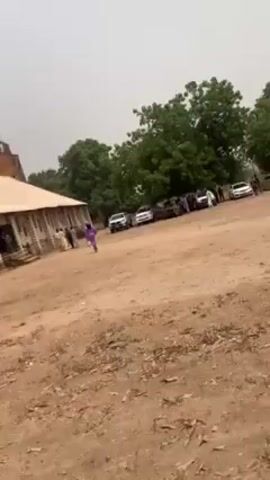 The video depicts an outdoor gathering in a rural area, possibly in Africa. In the background, a simple building with a pointed tower suggests a church or similar structure. Several vehicles are parked nearby.  The foreground shows a large open, dirt ground where people are milling about. A group of women in brightly colored, traditional clothing are drumming and dancing. Other individuals can be seen walking around, some wearing white garments. The overall scene seems to be part of a cultural or religious celebration.
