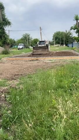 A Bobcat skid steer loader is positioned on a dirt construction site, with its front loader full of soil, appearing to be in the process of leveling or moving material. The loader is situated within a rectangular area defined by a border of some sort, possibly for a foundation or pathway. The ground is uneven and mostly bare earth, with areas of disturbed soil. Tall green grass and weeds are visible in the foreground and on the sides of the frame. In the background, a white SUV is parked on a paved surface, and utility poles and wires are visible. Trees and some buildings are also present in the distance, suggesting a semi-rural or suburban setting. The sky is overcast with clouds.