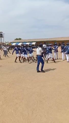 The video shows a marching band of students practicing or performing outdoors on a dirt field. The band members are wearing matching blue and white uniforms. Several adults in similar but distinct uniforms are observing and giving direction.  The band is marching and carrying flags. In the background, there is a low, long building with a blue and white awning. A water tower is visible in the distance. People can be seen walking and standing around the perimeter of the field. The sky is partly cloudy.