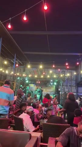 The video shows a crowded outdoor venue with a stage where a singer and a DJ are performing. People are seated at tables, socializing, and enjoying the entertainment. The stage is adorned with the logo of "Guinness Taste Naija's Favourite." Lighting is provided by strings of festive bulbs, and stage lights are visible.