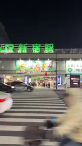 At night, a pedestrian crosswalk in front of the JR Shinjuku Station in Japan is the central focus. A steady flow of vehicles, including cars, vans, and motorbikes, pass by. People walk towards and away from the station entrance. The station is illuminated with bright lights, and advertisements are visible above the entrance. The green JR Shinjuku Station sign is prominently displayed on the building. The sky is dark.