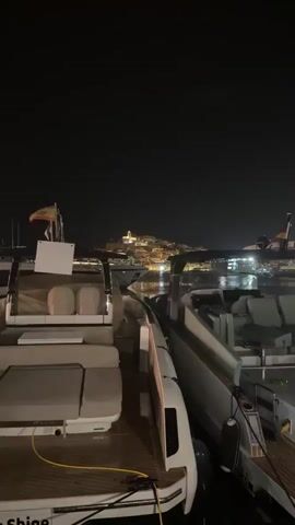 The video captures a nighttime scene at a marina with two luxury speedboats docked side-by-side in the foreground. The boats are sleek and modern, with plush seating and polished finishes. In the background, a town is illuminated by lights, perched on a hill overlooking the water. The lights of the town are reflected on the calm surface of the harbor. The sky is dark and clear. The overall atmosphere is one of luxury and tranquility.