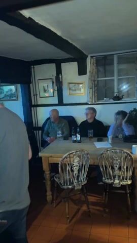 The video shows a scene from inside what appears to be a pub or tavern. There are several people present. Some are seated at wooden tables, drinking and observing, while others are dancing in a group near what looks like a band area, bathed in colorful stage lights. The room features a low ceiling with dark wooden beams. On the walls are framed pictures and a sign reading "PLEASE HEAD". The general atmosphere suggests a lively, social gathering with music and dancing.