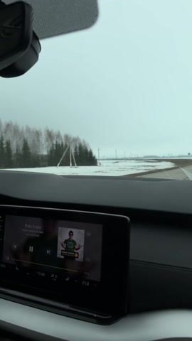 The video shows a car interior while driving on a road. The car's dashboard is visible, displaying a music player with a song by Макс Корж playing. The outdoor scenery visible through the windshield shows a flat landscape covered with patches of snow, power lines, and some trees in the distance under an overcast sky. The car is moving forward on the road. The scene appears to be taken during the daytime.