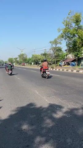 The video shows a sunny day on a paved road. Several people are riding motorcycles and scooters in the same direction. The road is wide and appears to be a highway or main road. There are trees and buildings on the side of the road, suggesting an urban or suburban environment. The sky is clear and blue. The camera is positioned at a low angle, possibly from another moving vehicle, giving a sense of forward motion.