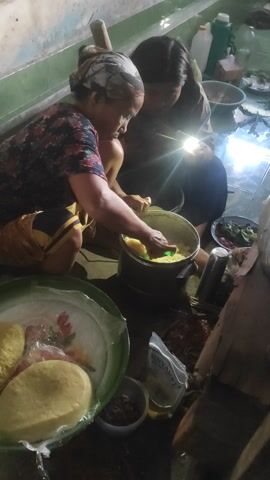 In a dimly lit interior, likely a home in Sakra, Indonesia, two individuals are engaged in food preparation. An older woman, wearing a floral patterned top and a headscarf, is seated and appears to be mashing or stirring a yellow substance in a pot with a spoon. Next to her, another person, possibly a younger individual, is leaning in, also focused on the task. A bright light source, possibly a flashlight or a camera flash, illuminates the pot and their hands, creating a strong glare.

The scene is cluttered with various objects: bowls of ingredients, a woven basket, and what looks like packaged goods, one with visible text that reads "Minyak" (Indonesian for oil). The setting suggests a rustic environment with wooden elements and a tiled floor. The presence of food items and the act of cooking imply a domestic activity, possibly preparing a meal for the family. The low light suggests it might be evening or early morning, and the absence of outdoor elements doesn't provide clues about the weather. The focus and close proximity of the individuals suggest a shared activity and possibly a sense of collaboration or familial connection. The overall mood is one of quiet concentration on the task at hand.