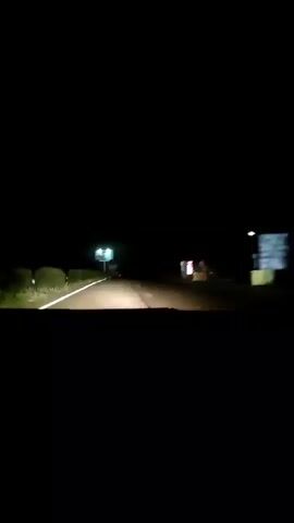 This is a first-person perspective video taken at night from inside a vehicle, likely on a highway. The headlights of the vehicle illuminate a dark road ahead. There are several signs visible in the distance. One prominent sign, illuminated by green lights, appears to be a large advertisement or informational billboard. Another smaller sign, possibly a directional marker or a business advertisement, is visible to the right, also illuminated. There are faint lights of other vehicles further down the road. The overall environment is dark and appears to be rural or semi-rural, with the road cutting through the darkness. There are no visible people in the frame, and the primary action is the vehicle moving forward.