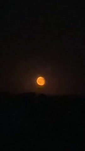 A video shows a bright orange moon set against a dark sky. The moon is partially obscured by a dark, silhouetted horizon, possibly trees or hills. There is no discernible movement in the video; it is a static shot of the moon in the night sky.