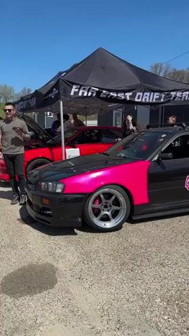 The video shows a parked, modified four-door car with a black and pink color scheme and the word "Sideway" in pink lettering on the rear quarter panel. The car has a roof rack and a large rear spoiler. It is parked near a tent labeled "Far East Drift Team" and a grey portable building. The sky is blue, and there are a few people standing around the car and tent. A red car can be seen parked behind the pink and black car, partially under the tent. The ground is paved.