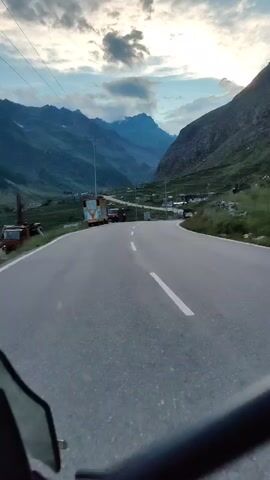 A POV shot from a motorcycle shows a paved road curving through a mountainous valley. The road has a white dashed line down the center and is bordered by green grassy slopes and rocky mountainsides. Several vehicles are visible on the road, including cars, a truck, and a utility vehicle. There are also a few buildings and structures, including what appears to be a small roadside shop or dwelling. The sky is partly cloudy with a soft, diffused light, suggesting it might be early morning or late evening. The overall impression is one of travel through a scenic, remote mountain landscape.