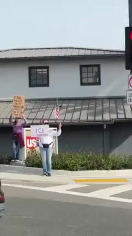 The video shows a series of scenes featuring people holding signs and protesting. In the first scene, four women stand near a usbank building, each holding a sign. The signs contain various messages, including one that says "Believe black lives matter". Another says "Stand With Ukraine". The next scene shows more protesters, some holding signs that say "Stand Up 4 Democracy". Another says "Abolish Trump". Later, a man is seen standing next to a woman with a dog, holding a sign. The sign says "Abolish ICE". An older man sits in a folding chair, seemingly observing the protest. Additional protestors hold signs saying "ICE = Border Patrol". Finally, the video shows protesters holding signs with messages such as "No ICE" and "Free Melani, Embrace Trump". The environment is an urban setting with buildings, streets, palm trees, and some greenery. Traffic can be heard throughout the video.