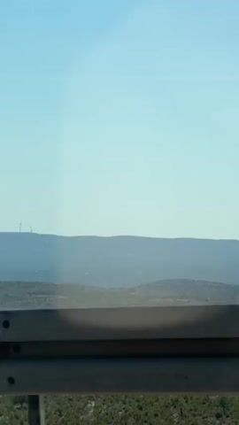 The video shows a landscape with rolling hills and a clear blue sky. In the distance, on top of a ridge, are several wind turbines. In the foreground, there is a dark metal railing. The camera appears to be moving, as the landscape shifts slightly throughout the short clip. There are no people or animals visible in the frame. The environment is dry and arid, with sparse vegetation. The lighting suggests it is daytime, likely with bright sunlight.