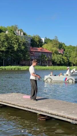 A young man stands on a wooden pier, fishing with a rod. In the background, on the water, a pedal boat shaped like a car, with BMW M colors on its side, is being pedaled by people. Another pedal boat shaped like a swan is also visible in the distance. The scene is set on a sunny day, with green trees and buildings on the shore.
