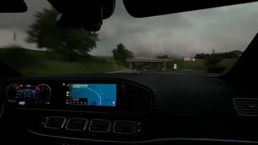 The video shows a first-person perspective from inside a car, driving on a highway. The dashboard is visible, with a digital speedometer on the left and a navigation screen in the center displaying a map with a blue route. Outside the car, the scenery consists of a road with trees and hills on either side. The sky is overcast and dark, suggesting cloudy or stormy weather. The car appears to be in motion, indicated by the blurred scenery and the car's instruments.