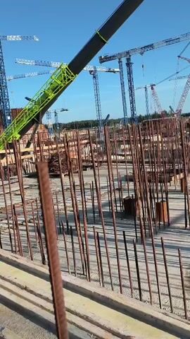 The video shows a construction site with numerous cranes and rebar. The camera is focused on a piece of rebar in the foreground, which is rusty and positioned vertically. In the background, several large blue and white cranes are visible, along with a bright green crane boom extending diagonally. The ground is covered with concrete and more rebar, indicating ongoing construction. The sky is clear and blue. The footage appears to be shot from an elevated perspective, possibly from another part of the construction site. There are no people visible in the frame.