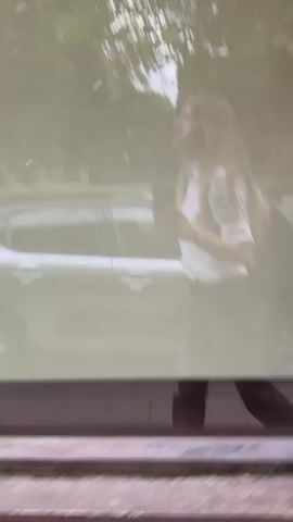 A woman with long blonde hair, wearing a white short-sleeved shirt and dark pants, walks past a car, holding a phone in her right hand. The scene appears to be a reflection on a window, with trees and buildings visible in the background. The woman is walking from right to left.
