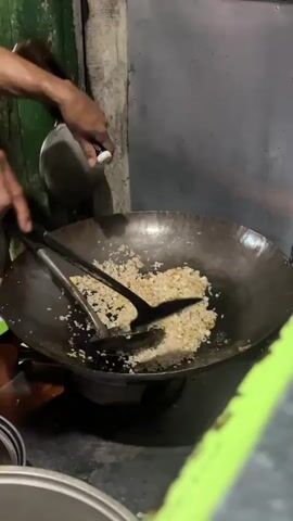 A close-up shot shows a pair of hands stirring rice in a wok over a flame. The rice is a light brown color and appears to be partially cooked. Two black cooking utensils are being used to stir the rice, one is a spatula and the other appears to be a ladle. The wok is dark gray and is sitting on a metal grate, which is positioned over a gas burner. The background is out of focus, but it appears to be a kitchen or food stall setting. There are some green and gray elements in the background, as well as some metal objects hanging on the wall. The overall lighting is dim, with the flame from the burner providing some illumination.