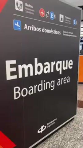 The video shows an interior view of an airport. Initially, there is a sign in Spanish and English that indicates "boarding area" and "domestic arrivals". A quick pan reveals a McDonald's restaurant and a view of the airport concourse. People are seen walking, some pulling luggage. Airport signs overhead indicate "check-in" and "domestic arrivals". The area appears clean and well-lit.