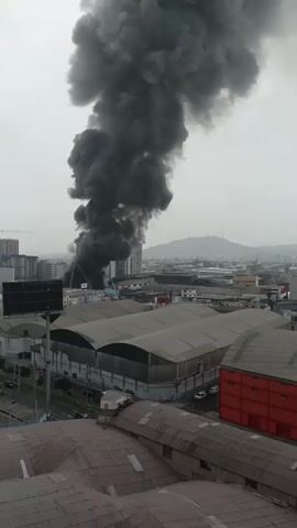 A plume of thick black smoke billows into the sky from a building in a city. Firefighting efforts are underway, with a ladder truck extending towards the smoke. Several other buildings and some industrial structures are visible in the foreground and background, under an overcast sky.