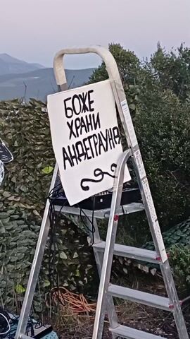 The video shows an outdoor musical performance by a band. A sign that reads "God Save the Underground" in a Slavic language, hangs on a ladder. A man plays a keyboard, a woman plays a tambourine, and two men play electric guitars. A drum set is visible in the background, along with amplifier and speakers. The band stands on a red patterned rug. The setting is a backyard type space with dense foliage, under cloudy, late-day light.