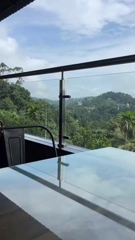 A stationary, eye-level outdoor shot shows a glass table and a glass and metal railing overlooking a lush green valley with rolling hills, dense foliage, and scattered white buildings under a partially cloudy sky.