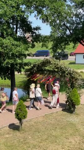 A group of people are walking under a large archway decorated with greenery and colorful ribbons. The archway is situated outdoors in a park-like setting with trees and a pond visible. People are casually dressed, suggesting a leisurely outing. The weather appears to be sunny and pleasant.