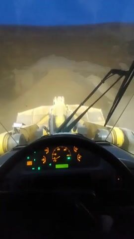 The video shows a first-person perspective from inside the cab of a large yellow front-end loader. The loader is moving through a pile of light-colored material, likely sand or gravel, at dusk or dawn as indicated by the dark sky. The headlights of the loader illuminate the material ahead. The dashboard is visible, showing various gauges and indicator lights, including a speedometer. The operator's hands are not visible, but the steering wheel suggests the machine is in motion. The view is primarily focused forward, with the loader's bucket and lift arms prominent in the scene.