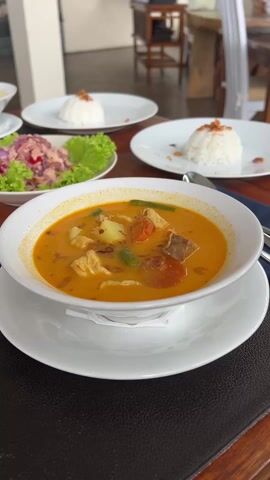 The video shows a table at a restaurant set for a meal. There are two bowls of orange soup with chunks of chicken and vegetables, two plates of white rice topped with fried onions, and a salad of chopped chicken or tuna, red onion, and lettuce leaves. There are also cutlery and blue napkins. The table is wooden and the setting appears to be relaxed and casual.