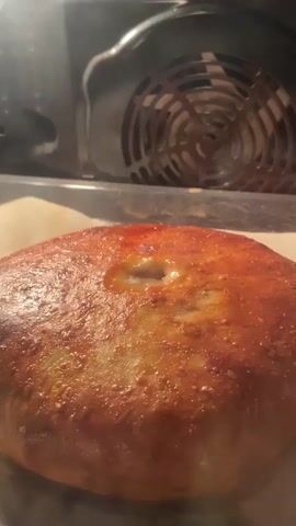 The video shows a close-up, slightly shaky shot of a golden-brown, round pastry with a hole in the center, baking in an oven. The pastry is sitting on what appears to be parchment paper on a baking sheet. Inside the hole, a small amount of liquid or filling is bubbling. The back of the oven is visible, with a circular fan or vent. The lighting is dim, likely from the oven light. The pastry is glistening and appears to be freshly baked.