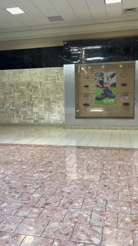 This is a first-person perspective video of walking through a seemingly empty shopping mall. The camera pans from left to right, showing the tiled floor with a reddish-brown marble pattern in the foreground and a lighter, beige tiled floor in the background. To the left of the frame is a store with a large display window showcasing shoes and a poster for Crocs. The walls of the mall are decorated with a textured, light-colored material. Further down the mall, to the right, more store windows and an empty display table with a plant are visible. The lighting appears to be fluorescent, typical of an indoor mall. There are no other people visible in the frame.
