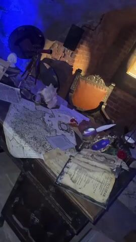 The video shows a room decorated in a historical style. A wooden desk is cluttered with old maps, books, a telescope, and other objects. A chair sits next to the desk. The walls are brick, and the ceiling is decorated with a pattern. The room is lit with a blue light. The camera moves, starting with a close-up of the desk and then panning up to the ceiling.