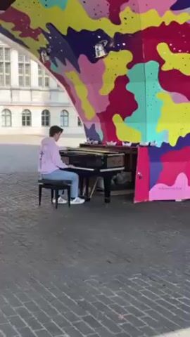 A young man wearing a light-purple hoodie, blue jeans, and white sneakers, is seated on a small black bench in front of a black upright piano, playing it. The piano is positioned in a public square with a brick-paved surface, under a brightly-colored structure. The structure appears to be a shelter with a rounded roof decorated with abstract shapes of various colors, including yellow, purple, pink, green, and light blue. There are small white paper boats placed on top of the piano, close to the colorful structure. On the right side of the piano is a painted door with a hashtag, and the building behind the structure is light-colored with many windows. The time of day appears to be late afternoon, with sunlight casting shadows on the brick pavement. The camera is positioned at eye level and remains stationary throughout the short clip.