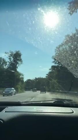 A first-person perspective video shows a view from the windshield of a car driving on a highway. The sun is shining brightly in the upper right corner of the frame, causing lens flare and glare on the windshield, which also appears to have some dirt or water spots. The highway ahead is clear with a few cars visible at a distance. Trees and greenery line both sides of the road. The dashboard of the car is visible at the bottom of the frame.