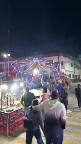 The video shows a night scene at what appears to be a carnival or fair. Several people are visible, some standing near a food stall labeled "La Ricosura," while others walk around. In the background, there are several carnival rides illuminated with colorful lights, including one that resembles an octopus and a very tall swing ride. The overall environment suggests a lively and festive atmosphere.