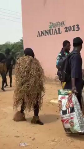 The video shows two people in costumes walking on a dirt road near a building with a sign that says "Tetfund Annual 2013". One person is wearing a suit made of brown straw, while the other has their body painted black and is wearing a blue head covering. A third person in a blue uniform walks past with a bag. The scene appears to be taking place outside in a rural or semi-rural area, with trees visible in the background.