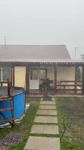 The video shows a backyard scene during a heavy rain. A yellow house with a brown metal roof and a covered porch is visible. A pathway of rectangular stone slabs leads from the foreground to the porch. To the left of the pathway is a large, blue, circular swimming pool covered with a black tarp, with a wooden ladder leaning against it. A small green structure with a wooden frame is next to the pool. The porch has wooden railings and furniture. The sky is overcast and the rain is visible as streaks falling from the sky. A large tree with green foliage is on the left side of the frame, obscuring part of the background. The overall environment appears to be rural or suburban.