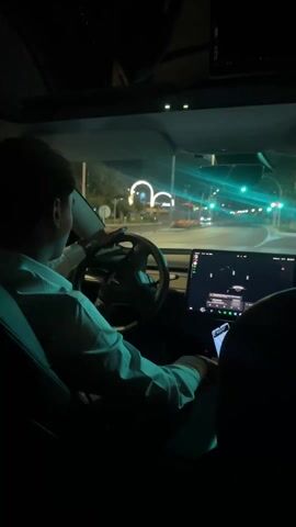 A man in a white shirt is driving a Tesla at night on a brightly lit street. The street has trees lining it and some decorative lights in the distance. He is holding his cell phone in his right hand, which has a map displayed on the screen. The central screen of the Tesla is on and playing music. He is wearing a watch on his left wrist.
